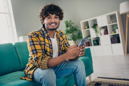 Photo of cheerful handsome guy sit sofa use smart phone chatting social network free time apartment indoorsの写真素材