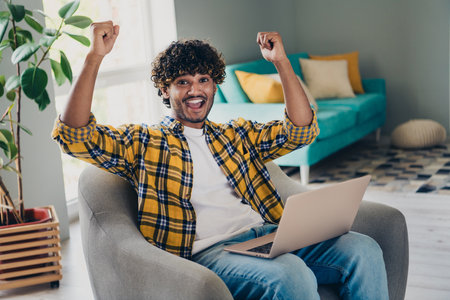 Photo of cheerful successful man sitting in comfortable armchair with raised hands watching football match weekend indoorsの写真素材