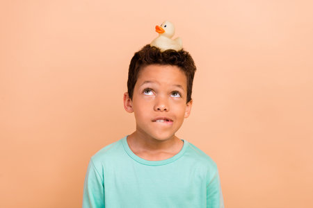 Photo of cute small boy wear stylish clothes bite lips look yellow rubber duck head nest isolated over beige color backgroundの写真素材