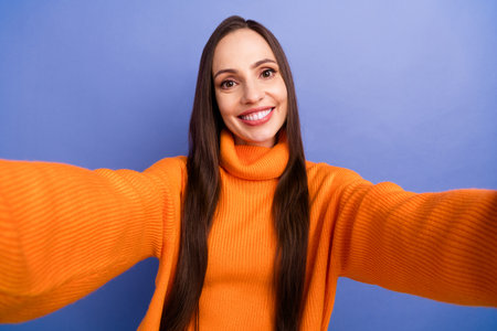 Self portrait photo of cheerful young lady in orange knitted turtleneck cozy atmosphere at home isolated on purple color backgroundの写真素材