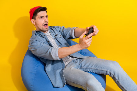 Photo of funky shocked guy wear denim jacket relaxing bean bag loosing game empty space isolated yellow color backgroundの写真素材