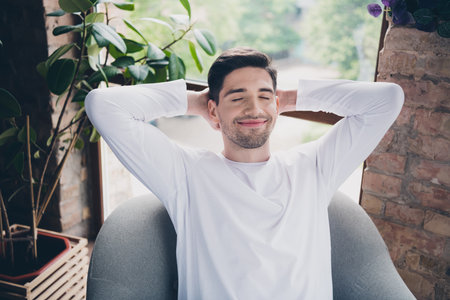 Photo of cheerful positive young guy wear white shirt smiling closed eyes relaxing arms behind head indoors apartment roomの写真素材