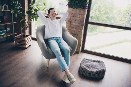 Photo of dreamy handsome young guy wear white shirt sitting armchair arms behind head indoors apartment roomの写真素材