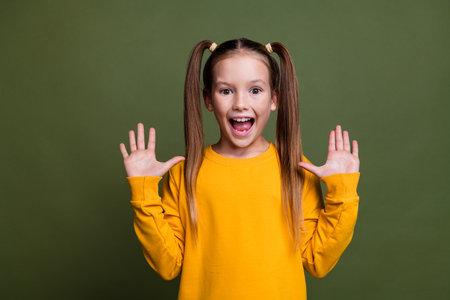 Photo of positive funny schoolgirl with ponytails wear yellow pullover showing palms open mouth isolated on khaki color backgroundの写真素材