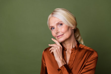 Portrait of cheerful pleasant person with blond hair wear stylish blouse touching chin near empty space isolated on khaki color backgroundの写真素材