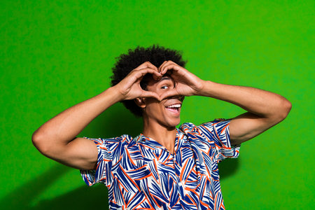 Portrait of attractive positive person happy young guy arms fingers demonstrate heart symbol eye isolated on green color backgroundの写真素材