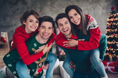 Portrait of four overjoyed peaceful people piggyback have fun enjoy fairy christmas time tree lights apartment indoorsの写真素材