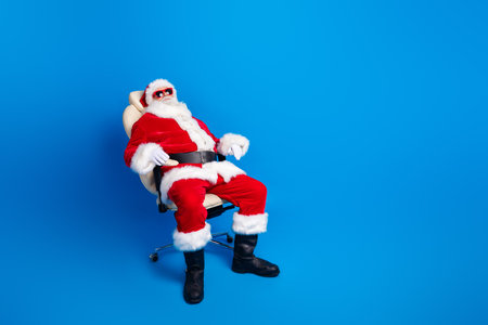 Full size photo of pensioner senior man sit relaxed office armchair wear trendy santa claus costume coat isolated on blue color backgroundの写真素材