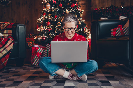 Full body photo of positive cheerful choosing shopping propositions mature aged lady sitting carpet at home with using netbook indoorsの写真素材