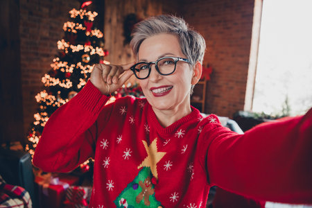 Selfie shooting of grandmother enjoying her outfit in red ugly sweater at home demonstrate atmosphere indoors touching glasses during xmasの写真素材