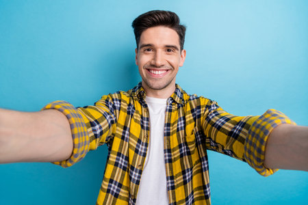 Photo of attractive cheerful guy wear yellow checkered shirt smiling recording video vlog isolated blue color backgroundの写真素材