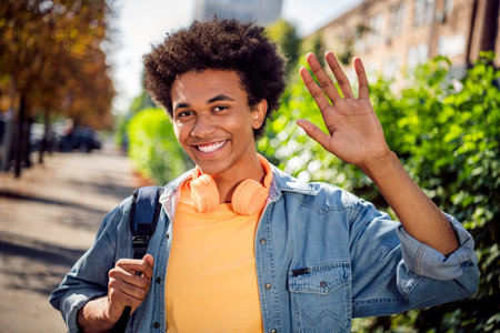 Photo of good mood cheerful guy dressed jeans shirt headphones waving arm palm hi outdoors urban city parkの写真素材