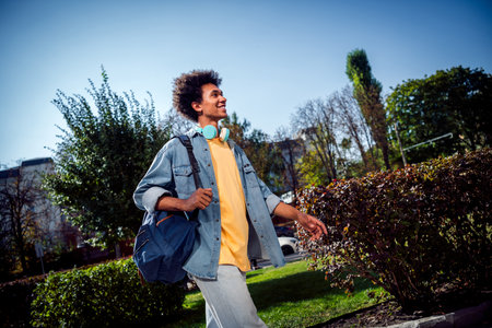 Photo of attractive cheerful guy dressed jeans shirt headphones walking university studies outdoors urban city parkの写真素材