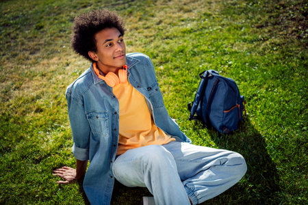 Portrait of satisfied relaxed guy with chevelure wear denim shirt jeans headphones sitting on grass after study with backpack outdoorsの写真素材