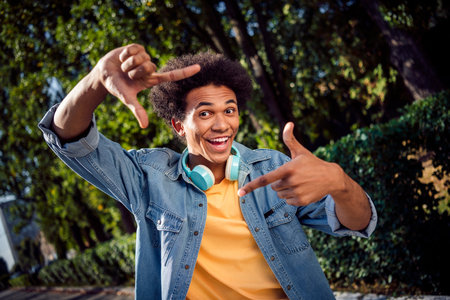 Photo of funky excited guy dressed jeans shirt headphones showing photo gesture outdoors urban city parkの写真素材