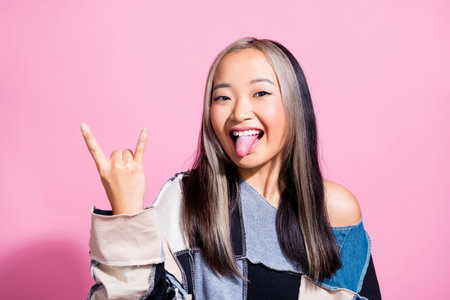 Photo of overjoyed indonesian girl tongue out demonstrate heavy metal symbol isolated on pink color backgroundの写真素材