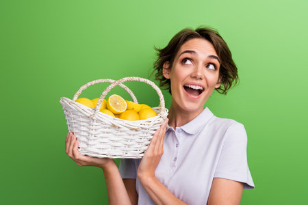 Photo of cheerful girl wear stylish outfit raise basket with fresh tasty fruits look up empty space isolated on green color backgroundの写真素材