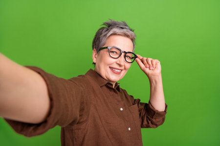 Photo of good mood smart woman with short hairdo wear brown shirt touching eyewear doing selfie isolated on green color backgroundの写真素材