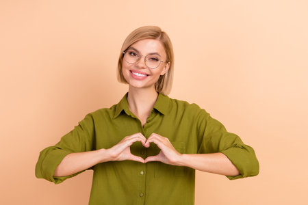 Portrait of toothy beaming girl with straight hair wear khaki shirt in eyewear showing heart on chest isolated on pastel color backgroundの写真素材