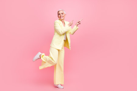Full size photo of cheerful woman wear yellow suit stand on one feet indicating at sale empty space isolated on pink color backgroundの写真素材