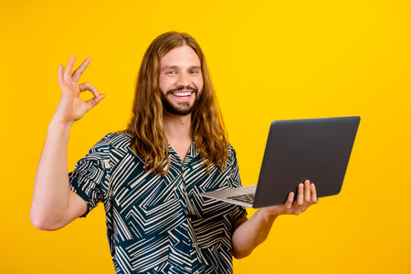 Photo of cheerful confident man dressed print shirt typing modern gadget okey gesture isolated yellow color backgroundの写真素材
