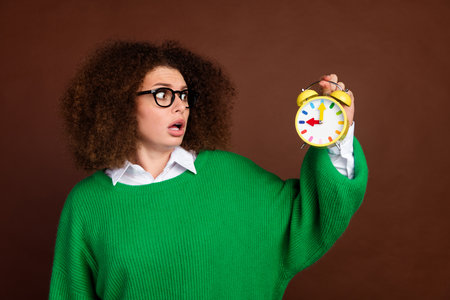 Portrait of worried speechless girl open mouth look hand hold alarm clock isolated on brown color...
