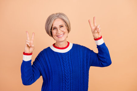 Photo of toothy beaming woman with gray hair dressed knitwear warm sweater showing v-sign symbol isolated on beige color backgroundの写真素材