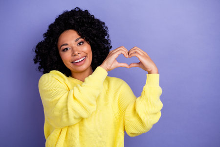Photo of toothy beaming woman with afro hairdo dressed yellow sweater fingers showing heart symbol isolated on violet color backgroundの写真素材