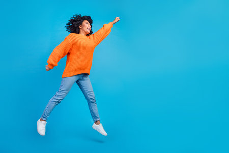 Full size photo of pretty young girl jumping superhero flying dressed stylish knitted orange outfit isolated on blue color backgroundの写真素材