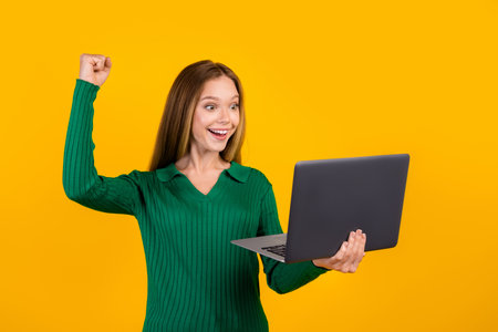 Photo of young girl teenager successfully done examination test at college raised fist up hold laptop isolated on yellow color backgroundの写真素材