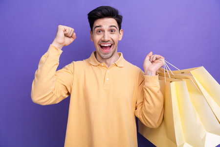 Photo portrait of attractive young man fist celebrate big sales shopping dressed stylish yellow clothes isolated on purple color backgroundの写真素材