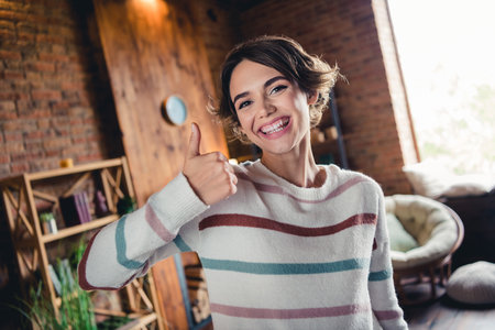 Photo of cheerful lovely girl toothy smile hand finger demonstrate thumb up approval loft apartment insideの写真素材