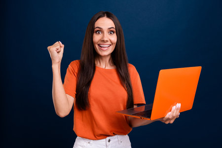 Photo of pretty lucky woman dressed orange t-shirt winning a laptop isolated dark blue color backgroundの写真素材