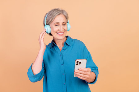 Photo of good mood senior woman with bob hairstyle listen playlist in headphones look at smartphone isolated on pastel color backgroundの写真素材
