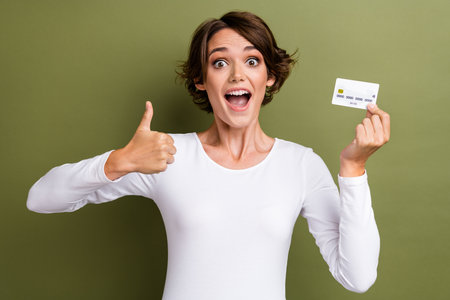 Photo portrait of pretty young girl hold credit card excited thumb up dressed stylish white outfit isolated on khaki color backgroundの写真素材