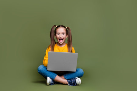 Full length photo of impressed overjoyed kid with ponytails hair sit on floor with laptop on legs isolated on khaki color backgroundの写真素材