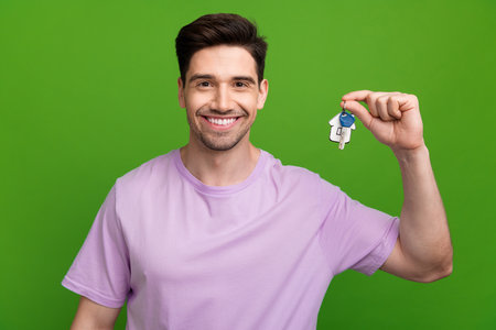 Photo of toothy beaming man with brunet hair dressed purple t-shirt holding keys from new apartments isolated on green color backgroundの写真素材