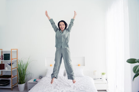 Photo portrait of attractive young woman jump bed raise hands cheerful dressed stylish gray pajama isolated on white modern interior designの写真素材