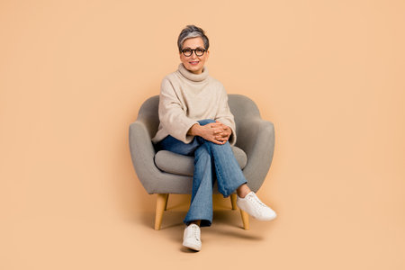 Full length photo of satisfied psychotherapist experienced mature age woman spend time on therapy sit armchair over beige color backgroundの写真素材