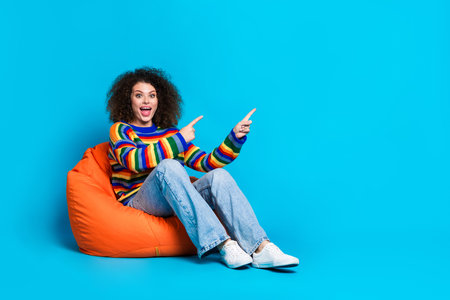 Full body photo of attractive young woman beanbag point empty space dressed stylish rainbow print clothes isolated on blue color backgroundの写真素材