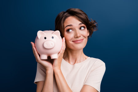 Photo portrait of pretty young girl look admire hold piggy account bank wear trendy white outfit isolated on dark blue color backgroundの写真素材