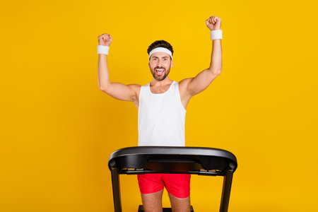 Photo of funny lucky guy sportswear rising fists winning treadmill race isolated yellow color...