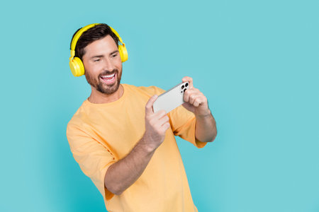 Photo of young funny freelancer guy makes money in gaming mobile apps watching videos wear headphones isolated on cyan color backgroundの写真素材