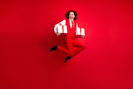Full length size photo of crazy surprised teenager boy wear formal suit jump with two wrapped packages isolated on red color backgroundの写真素材