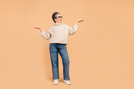 Full body portrait of positive grandmother look arms hold demonstrate empty space isolated on beige color backgroundの写真素材