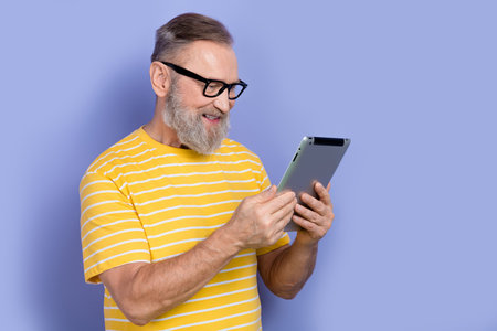 Photo of smart cheerful man with gray beard dressed striped t-shirt in eyewear read news on tablet...