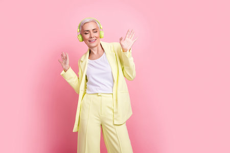 Photo of woman senior age in yellow suit dancing enjoying wireless headphones isolated on pink color backgroundの写真素材