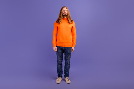 Full length photo of young long haired blond male model in orange basic sweater combination with jeans isolated on purple color backgroundの写真素材