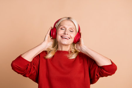 Photo of cheerful dreamy lady dressed red pullover closed eyes enjoying songs headphones isolated beige color backgroundの写真素材