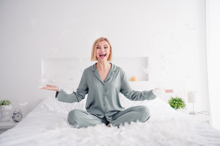 Photo portrait of lovely blonde young lady sit bed excited flying feather wear trendy gray sleepwear comfortable bedroom daytimeの写真素材